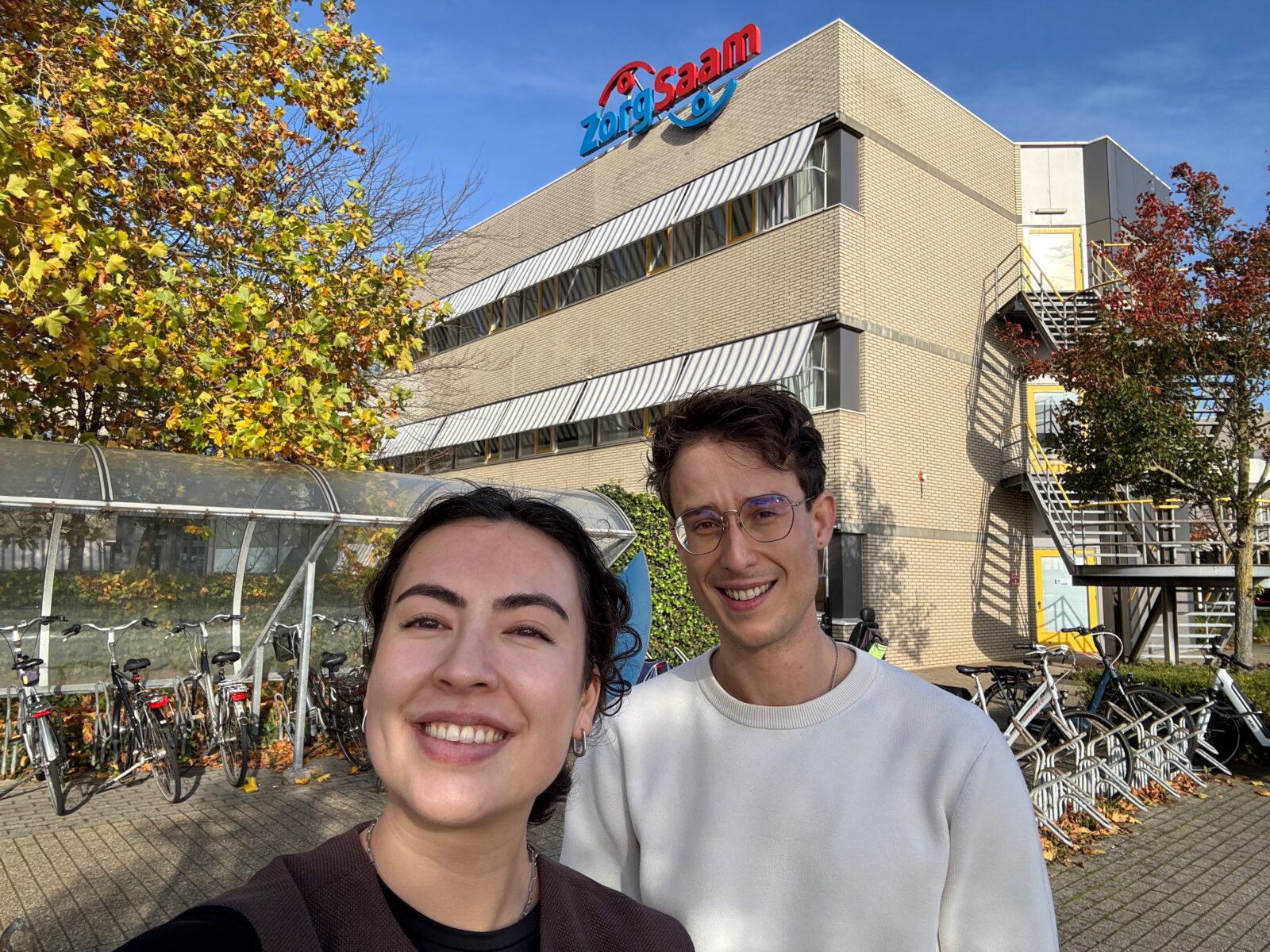 Selfie van trainees Selina en Thom voor de ingang van opdrachtgever ZorgSaam